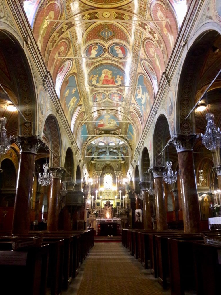 An old church aisle with marble columns and arches to either side. The ceiling is brightly painted with saints and Christian symbols