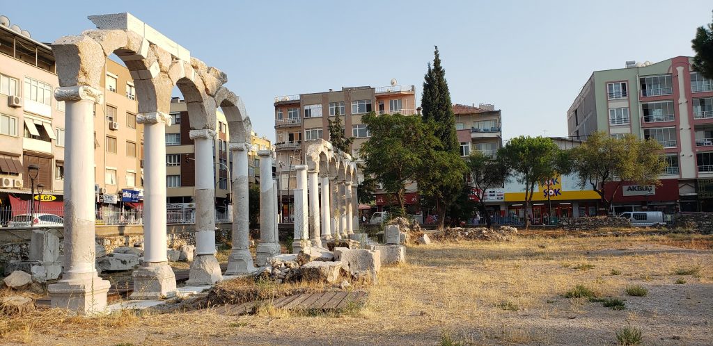 Thyatira: Seven Churches of Revelation. A flat grassy area, surrounded by buildings, contains the remains of a colonnade, some of the upper portions still intact 
