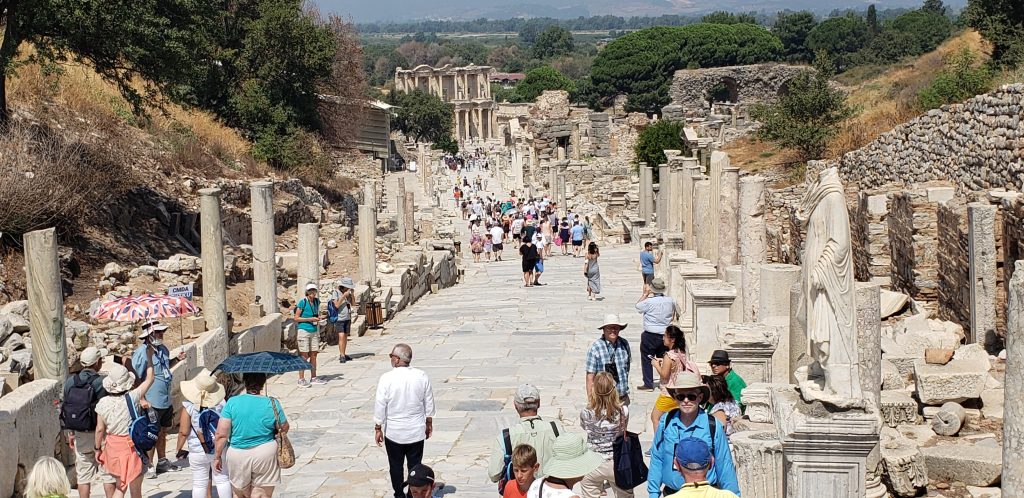 Modern tourists walk down an ancient road, paved with large slabs. The library facade is at the end of the road, far downhill, while the remains of columns stand on either side of the road
