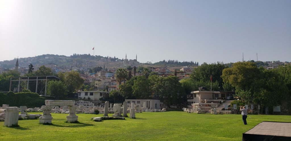 Smyrna: one of the Seven Churches of Revelation. A grassy plateau holds columns in the remains of the agora, with the city of Izmir on the hill behind. A large flag stands on top of the old citadel.