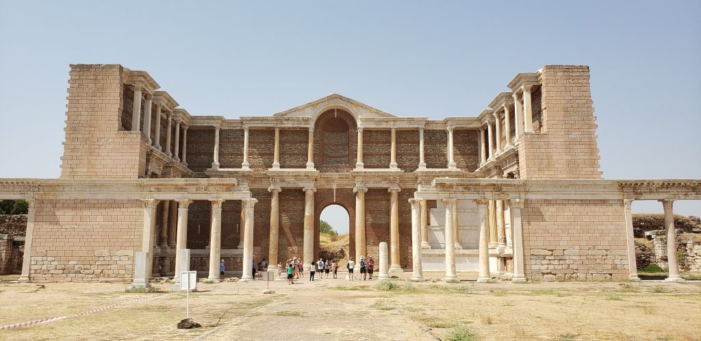 Sardis: one of the Seven Churches of Revelation. A two-level colonnaded building stands reconstructed, with three of its walls built to full height, and tourists walking within the building show that it is large.