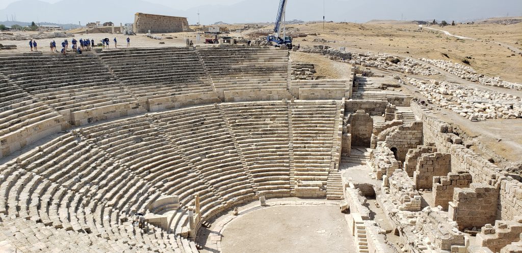 Laodicea: one of the Seven Churches of Revelation. Tourists (tiny dots in the distance) stand on the same level as our perspective. A large semi-circular theater leads downwards and the remains of a stage stand facing the seating