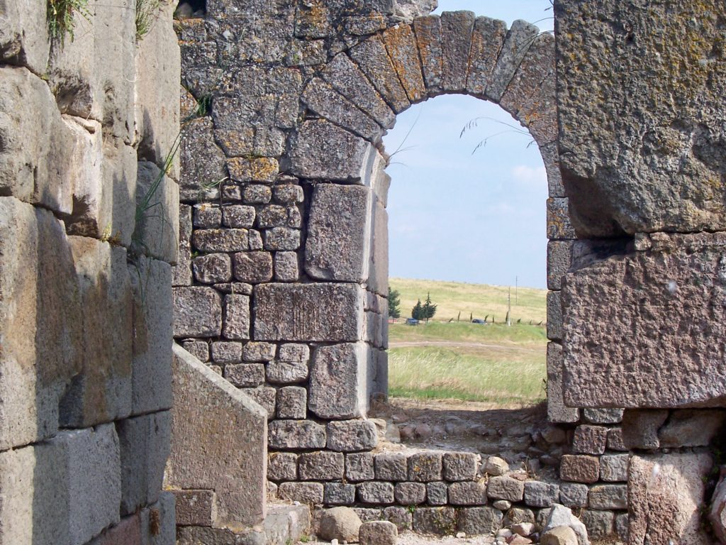 Remains of an old stone wall made of different-sized blocks, with the arched window still intact. The grassy hill behind is visible through the window.