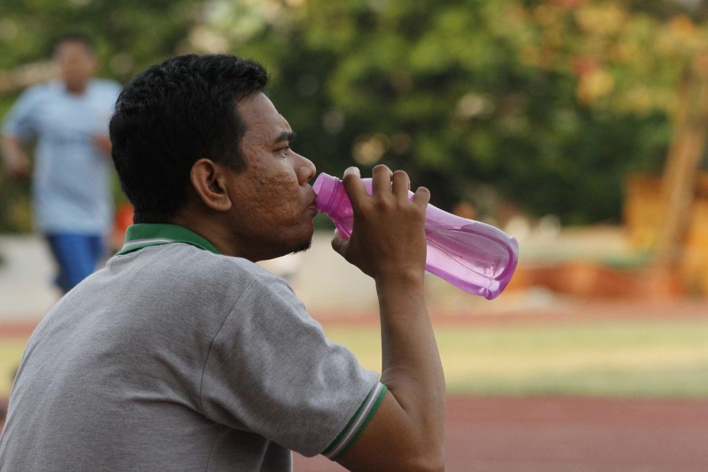 A dark-skinned man with black hair drinks from a reusable water bottle with trees in the background.