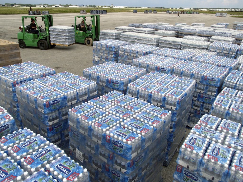 Huge packs of plastic bottled water by the side of an airport runway, with forklift operators loading and moving the pallets.
