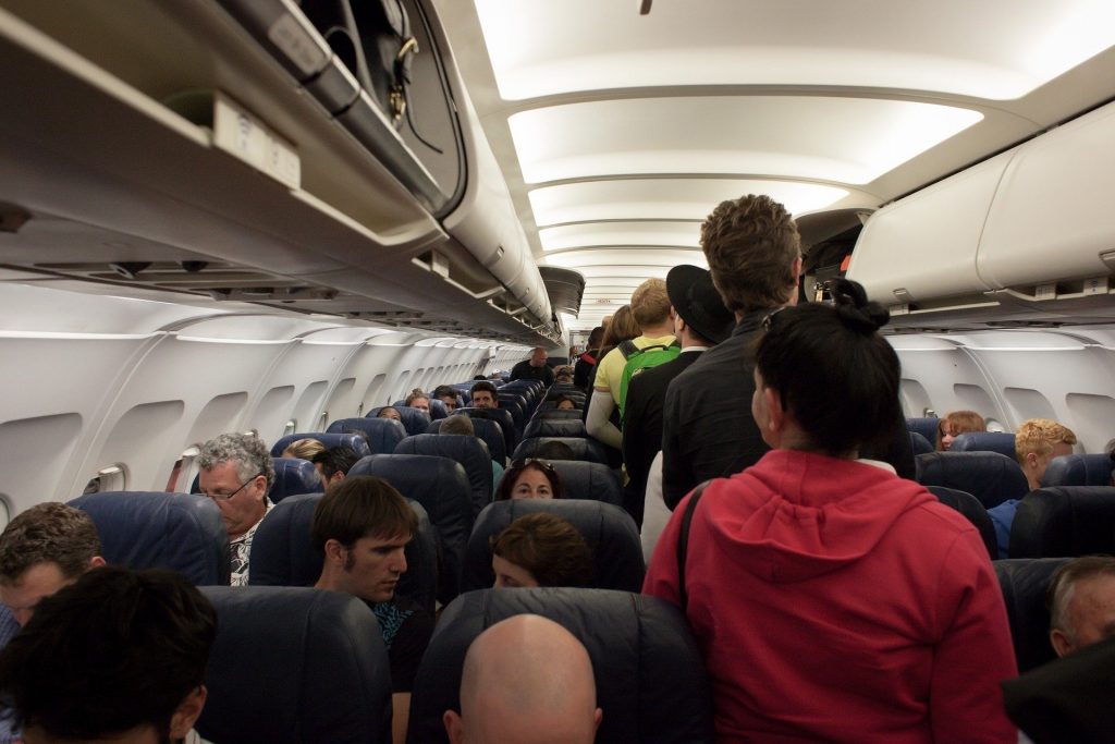 Passengers board a medium-sized aircraft (pre-Covid; no one is masked). There are three seats on either side of the single aisle and some overhead bins are open. Many passengers queue in the aisle. We will talk about ethical travel and airlines.