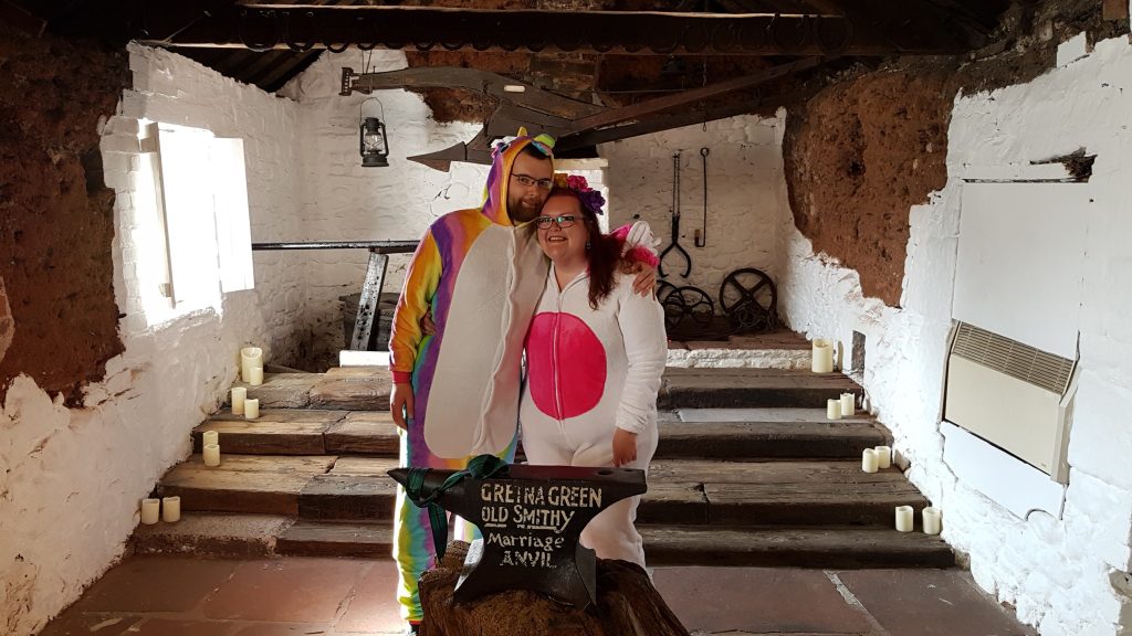 A couple stands in the Old Blacksmith's Shop in front of an anvil reading "Gretna Green Old Smithy: Marriage Anvil". They are wearing unicorn onesies.