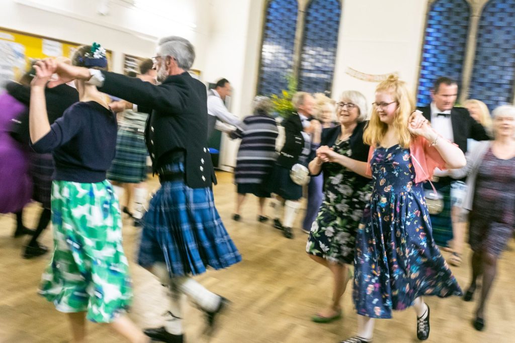 Happy wedding guests - in kilts, pretty dresses, and (most importantly) sensible shoes - dancing the ceilidh dance the Gay Gordons, holding hands above their shoulders.