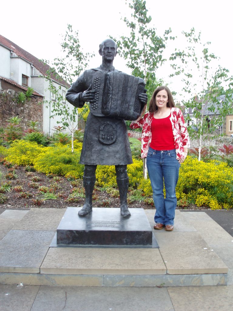 A picture of our founder Meg, with the dark metal statue of Sir Jimmy, 20th century proponent of ceilidh music.